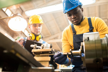 Serious concentrated African-American factory engineer in hardhat adjusting milling machine and...