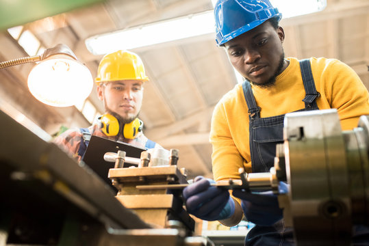 Serious concentrated African-American factory engineer in hardhat adjusting milling machine and joining detail while skilled inspector examining his work and controlling production process