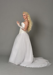full length portrait of blonde  girl wearing white fantasy gown. standing pose in side profile, grey studio background.
