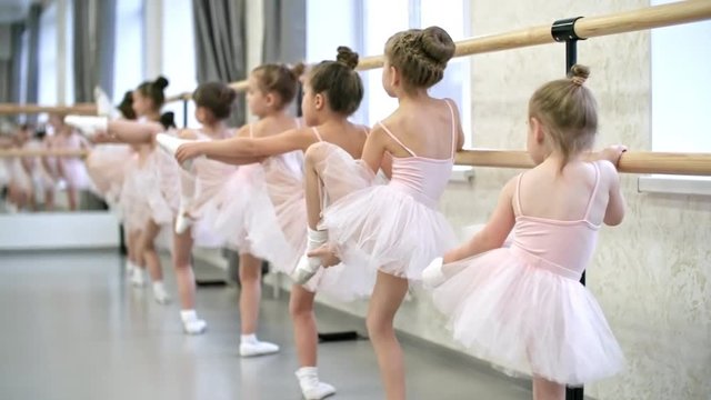 Rear view of group of little girls using ballet barre when doing leg stretching exercises in dance studio