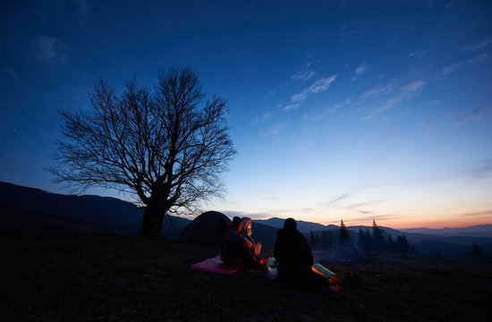 Camping site in mountains at dawn. Group of three young tourists sitting in front of tent and meeting early morning watching sky red from rising on horizon sun. Tourism and traveling concept.