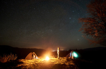 Night camping in mountains. Young tourist woman standing between two illuminated tents at burning campfire near big tree, watching sky with lot of bright sparkling stars. Tourism and travel concept