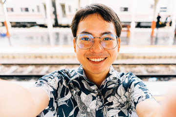 Asian young man holding camera and making selfie and smiling at train station.