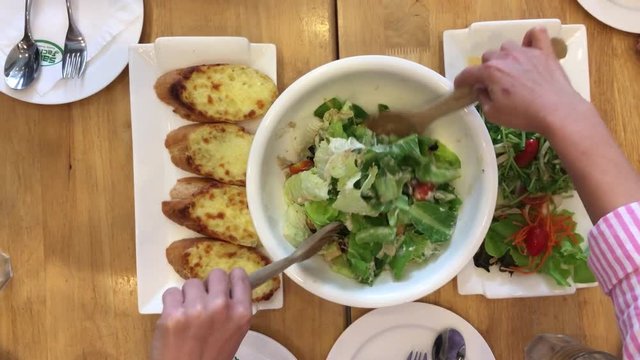Hand of woman mixing delicious superfood caesar salad ingredients with wooden spoons on the table at restaurant with garlic bread and food set. Cooking concept. Top view.