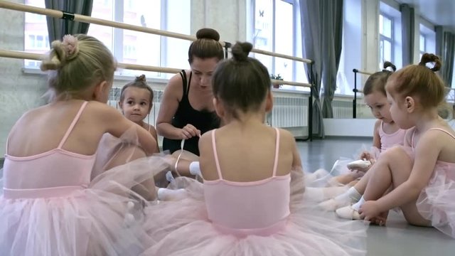 Group of little girls sitting in circle on the floor, their female teacher helping one of them tie ribbons of ballet shoes