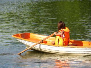 A girl in a life jacket floats on a rowing boat on a summer lake. Young woman rowing with oars,...