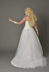 full length portrait of blonde girl wearing white gown, standing pose with back to the camera. grey studio background.