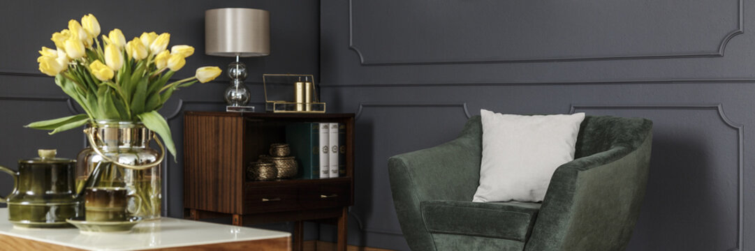 White pillow on a velvet green armchair and a wooden cabinet by a gray wall with molding in a classy living room interior. Real photo.