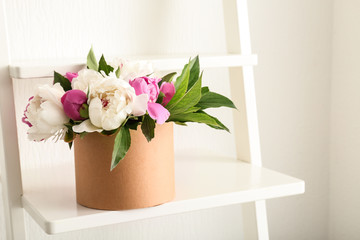 Box with beautiful peony flowers on shelf near white wall