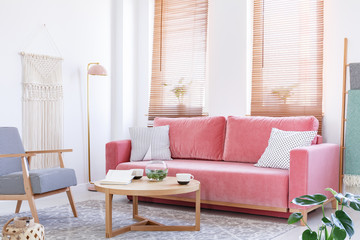 Patterned armchair next to pink sofa in bright flat interior with wooden table and blinds. Real photo