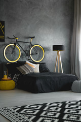 Bike above black bed against concrete wall in modern bedroom interior with patterned rug. Real photo