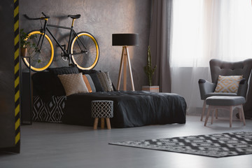 Bike above black bed in dark bedroom interior with patterned stool and armchair. Real photo