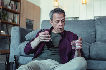 Glass of vodka. Mature badly looking man with facial wrinkles sitting with glass of vodka being alcohol addicted