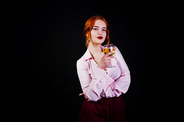 Gorgeous red haired girl in pink blouse and red skirt with glass of wine at hand isolated on black.