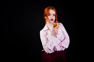 Gorgeous red haired girl in pink blouse and red skirt with glass of wine at hand isolated on black.