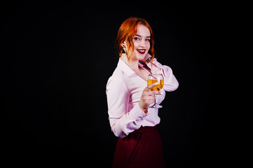 Gorgeous red haired girl in pink blouse and red skirt with glass of wine at hand isolated on black.