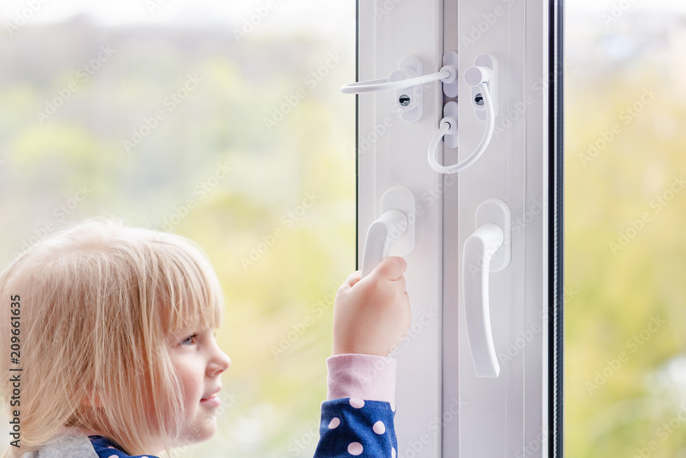 Wall mural Little cute toddler girl trying to open window in apartment at high-tower building. Children window protection lock. Cable safety guard prevent opening window by child. Prevention of falling accident