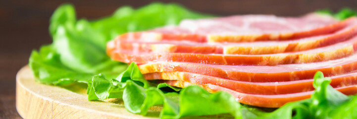 Sliced ham with fresh green lettuce leaves on a round cutting board. Meat products on a brown wooden table.. Banner