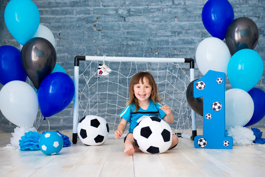 Little cheerful child dressed in sports clothes sitting on the floor near a football goal, looking at a big soccer ball. The first year, number one