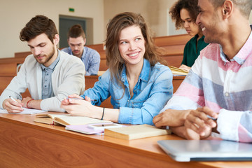 Fototapeta premium Cheerful positive beautiful student girl in denim shirt smiling while talking groupmate during break in university classroom, happy friends sitting at desk with books