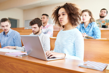 Obraz premium Pensive concentrated young multiethnic students sitting at university desk and attentively listening to lecturer, serious African-American girl with Afro hairstyle typing on laptop