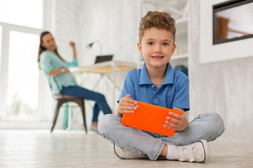 Near mother. Cute smart boy smiling broadly while playing online games sitting near working mother