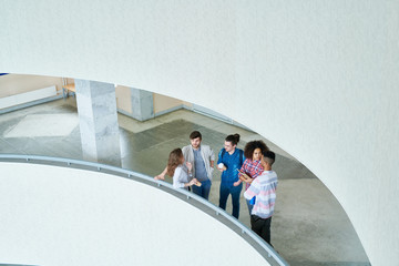 Top view of confident interracial student friends in casual clothing standing at railing in university hall and chatting while waiting to start class