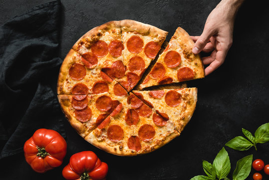 Hand picking slice of pepperoni pizza. Pizza party. Hot italian pepperoni pizza on black concrete background, top view