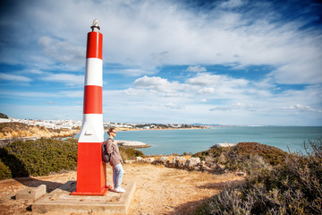 A girl traveler stands by the lighthouse atop a hill and looks at the ocean, the Algarve, Portugal, a popular destination for travel in Europe