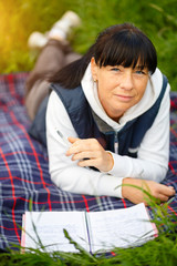 Cute brunette woman 35-40 years old lying on park grass with notebook on rug with pen in hand on nature, beautiful summer sunset evening. Education concept