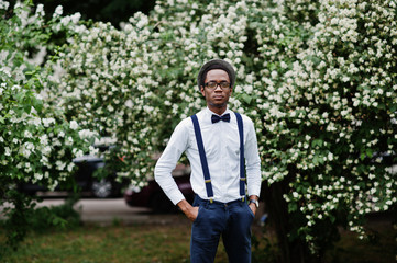 Stylish african american business man at pants with suspender and shirt with bow tie, hat and glasses posed outdoor.