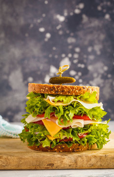 Big sandwich with meat (porshutto), cheese and fresh vegetables on a wooden board, dark background.