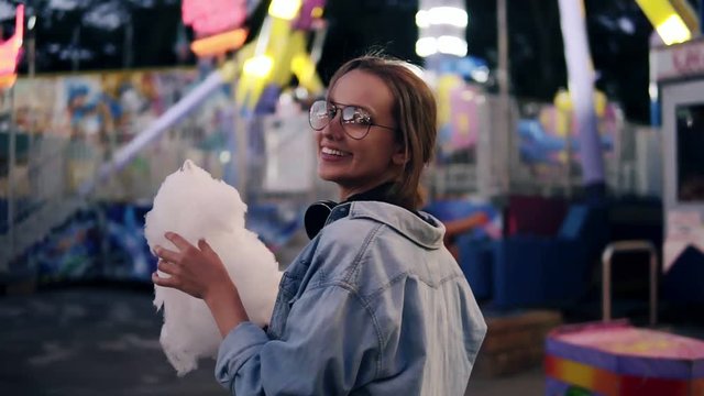 Traking footage of a young blonde girl walking in the amusment park, eating white candy cotton. Looking over her shoulder and smiling to the camera. Evening dusk