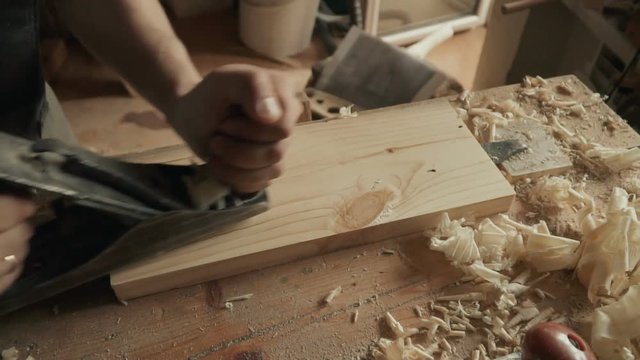 Slow motion - skilled craftsman carpenter whittles workpiece of wooden board with jack-plane in a workshop. Making handmade furniture.