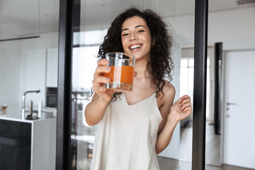 Photo of gorgeous brunette curly woman wearing silk leisure clothing smiling, while showing glass...