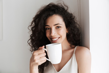 Portrait closeup of gorgeous happy woman with long curly hair smiling at camera, and drinking tea...