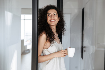 Photo of adorable curly woman with long dark hair smiling and looking aside, while standing near glass door in house with cup of tea