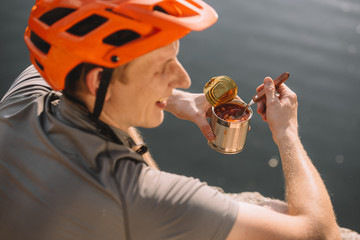 handsome young traveller in helmet eating canned food in front of water