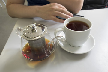 Glass teapot and white cup with tea on a light table and hands of a man with a mobile phone in the background