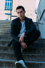 Sexy man posing sits on the steps near railing. Handsome young man in stylish black clothes and white shoes posing near a railing