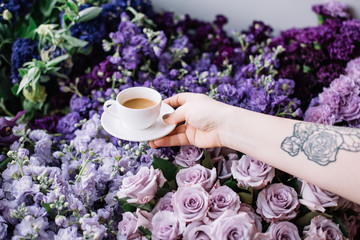 VORONEZH/RUSSIA-03.05.2018: Woman with a tattooed hand holding a cup of fresh morning espresso coffee in a white cup with purple colored flowers on the background (roses, carnations, mattiola, tulips)