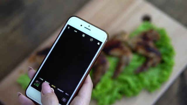 A woman takes pictures of food on her phone (smartphone). Fried chicken wings on a wooden board in tomato and dill.