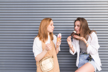 Portrait of two attractive girls of girlfriends in stylish light clothes, standing against a background of gray walls and eating ice cream. Cute, fashionable girls eat ice cream on a gray background