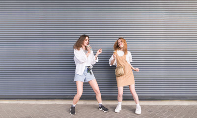 Two young girlfriends in stylish clothes dance on a gray background. Portrait of two cheerful girls on the background wall.