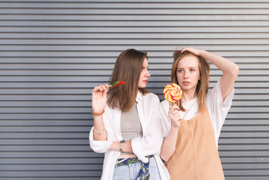 Two stylish young women in a light summer dress posing on a gray background with candies. Portrait of attractive girls with lollipops on the background of a dark wall.