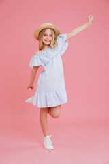 Full length portrait of cheerful kid summer girl 8-10 wearing blue dress and straw hat posing at camera with beautiful smile, isolated over pink background