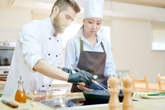 Two professional chefs working in modern restaurant kitchen standing at wooden table and cooking meat together, copy space