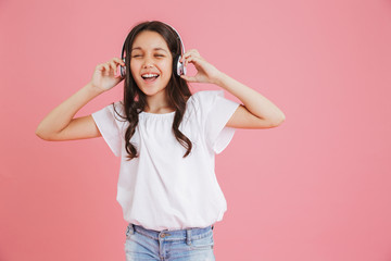 Obraz na płótnie Canvas Joyful young girl 8-10 in casual clothing singing with closed eyes while listening to music via wireless headphones, isolated over pink background