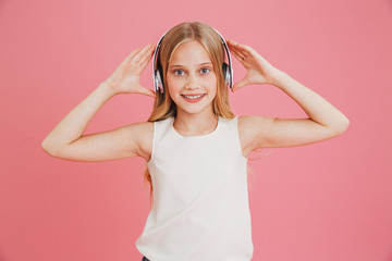 Beautiful teen girl 8-10 in casual clothing listening to music and touching wireless headphones at head, isolated over pink background