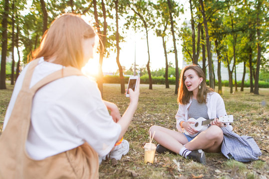 Girl takes the girl's ukulele at the camera. Shooting a cover on ukulele. The two girls sit in the park, one plays ukulele, the other takes it to the camera of the smartphone. Musical concept.